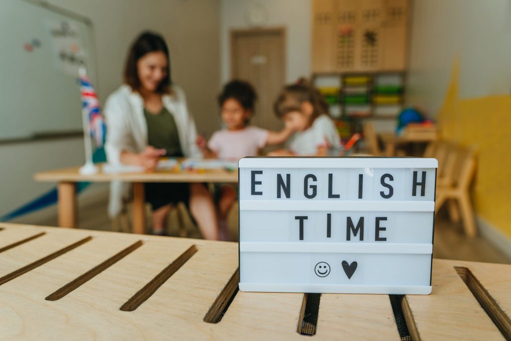 rotulo Hora del Idioma Inglés con maestra y niños en segundo plano